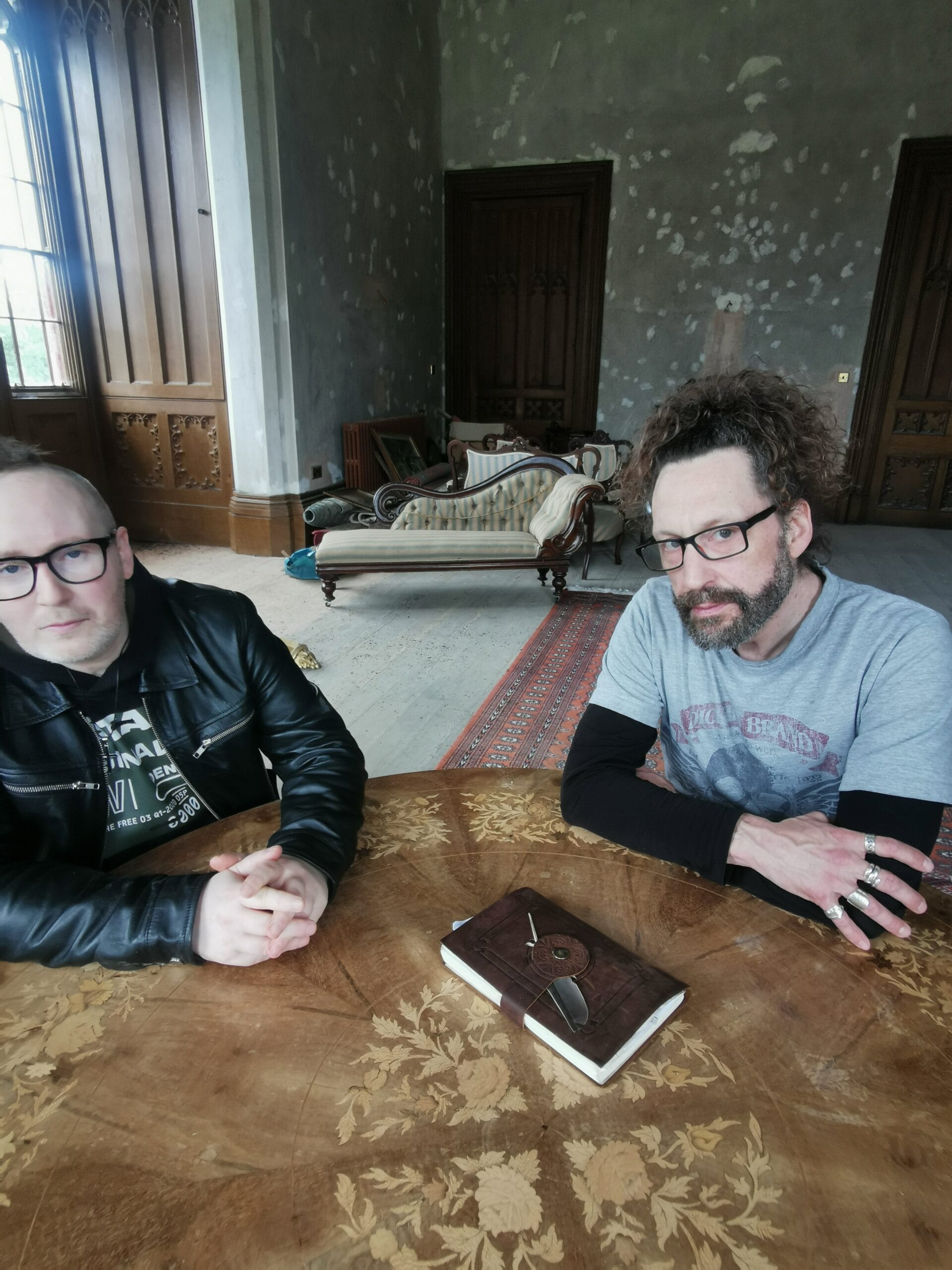Simon and Kelly sitting at a table with a leather-bound book in castle gothic-style room.