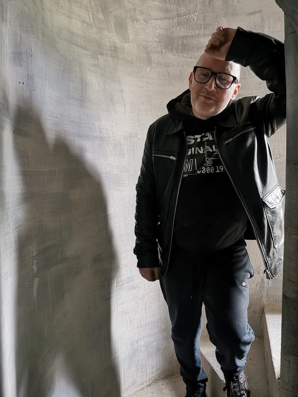 Simon Collins in leather jacket and glasses stands in a concrete stairwell, looking at the camera.