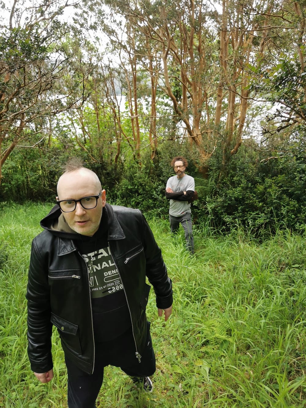 Simon and Kelly in castle grounds forest area, Simon in a leather jacket, Kelly with arms crossed.